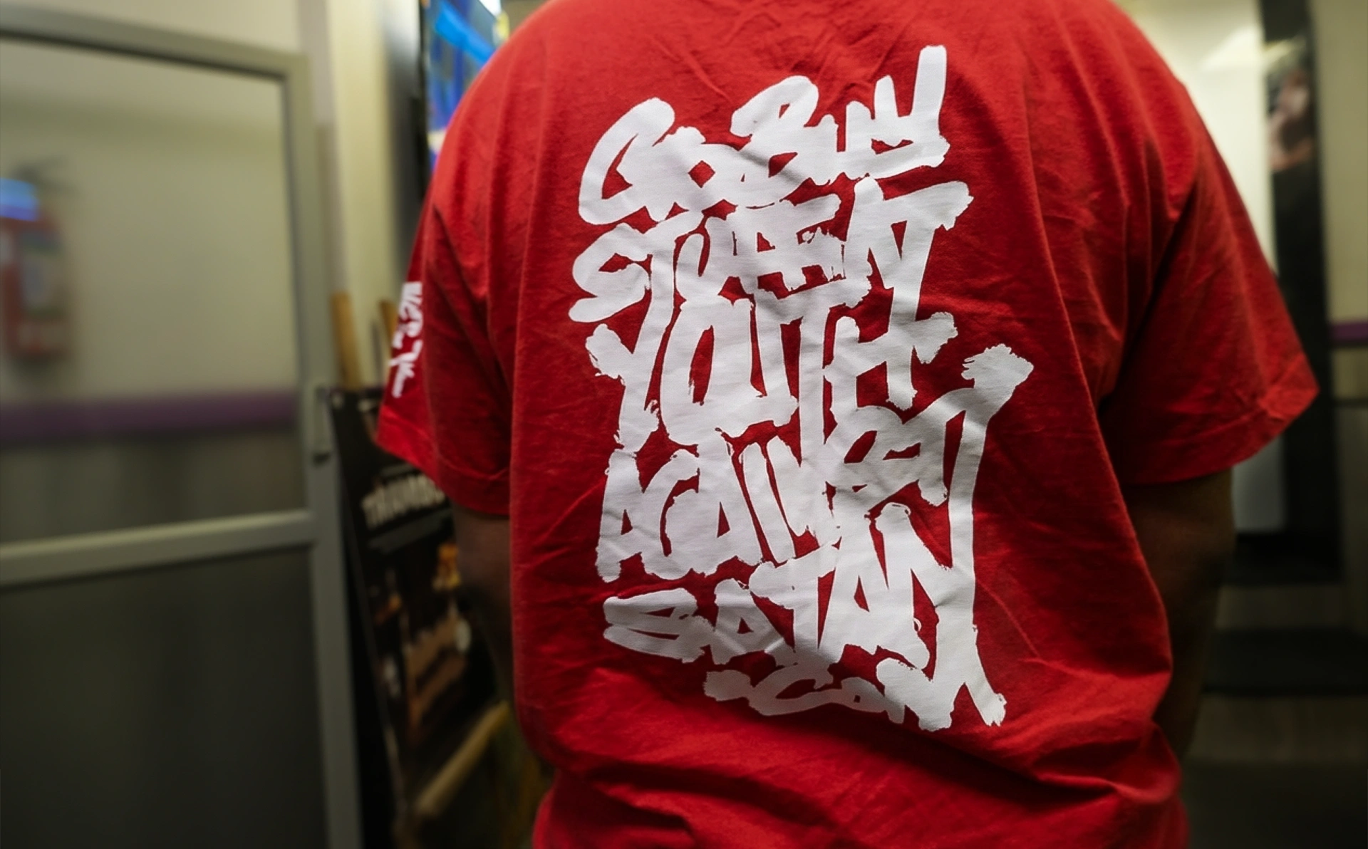 Backside of a red roundneck tee with a large white sticker print of graffiti-style text that reads “go buy stuff at youthagainstsatan.com”. On the left sleeve is seen a slightly out-of-focus portrait of Lex Walton made using a few white brush strokes.