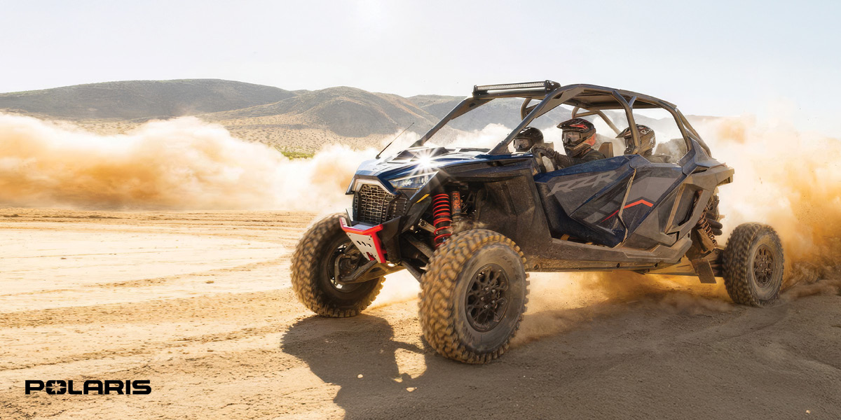 Blue Polaris off-road vehicle kicking up dust while driving on a sandy terrain with three people wearing helmets inside.