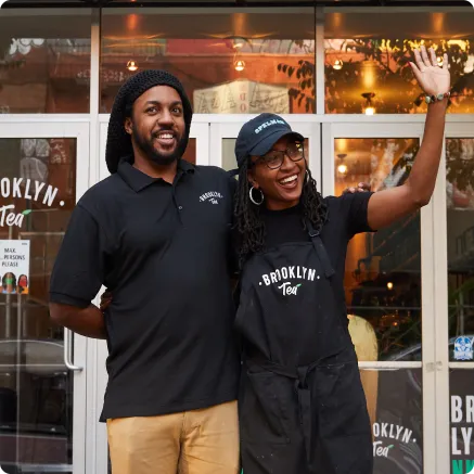 Two smiling people wearing Brooklyn Tea uniforms standing in front of a storefront, one waving.