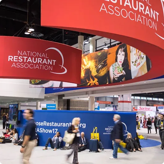 Convention scene with people walking and sitting near National Restaurant Association banners and booths.
