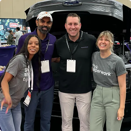Four smiling people standing in front of an open Ford vehicle hood at an indoor event.