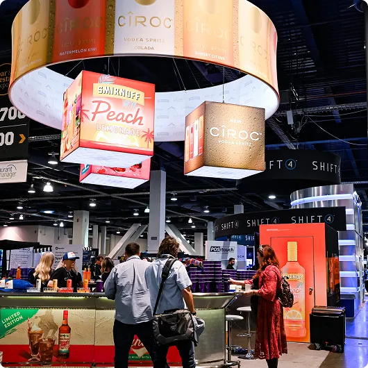 Trade show booth featuring illuminated hanging signs for Smirnoff Peach Lemonade and Ciroc vodka, with people engaging at the counter.