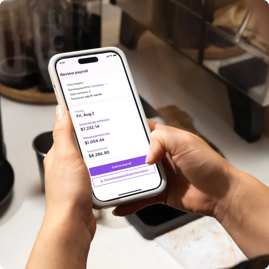Person holding a smartphone displaying a payroll review screen with payroll details and submit and download buttons.