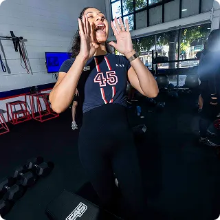 Woman in workout attire shouting or cheering in a gym setting with exercise equipment in the background.