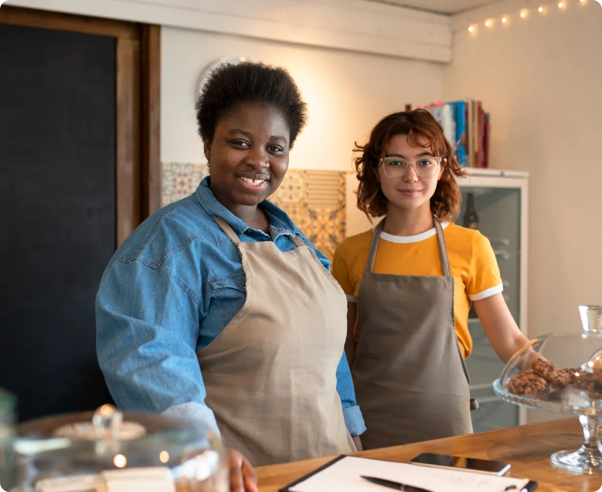 Two smiling cafe workers wearing aprons standing behind a wooden counter with baked cookies under a glass dome.