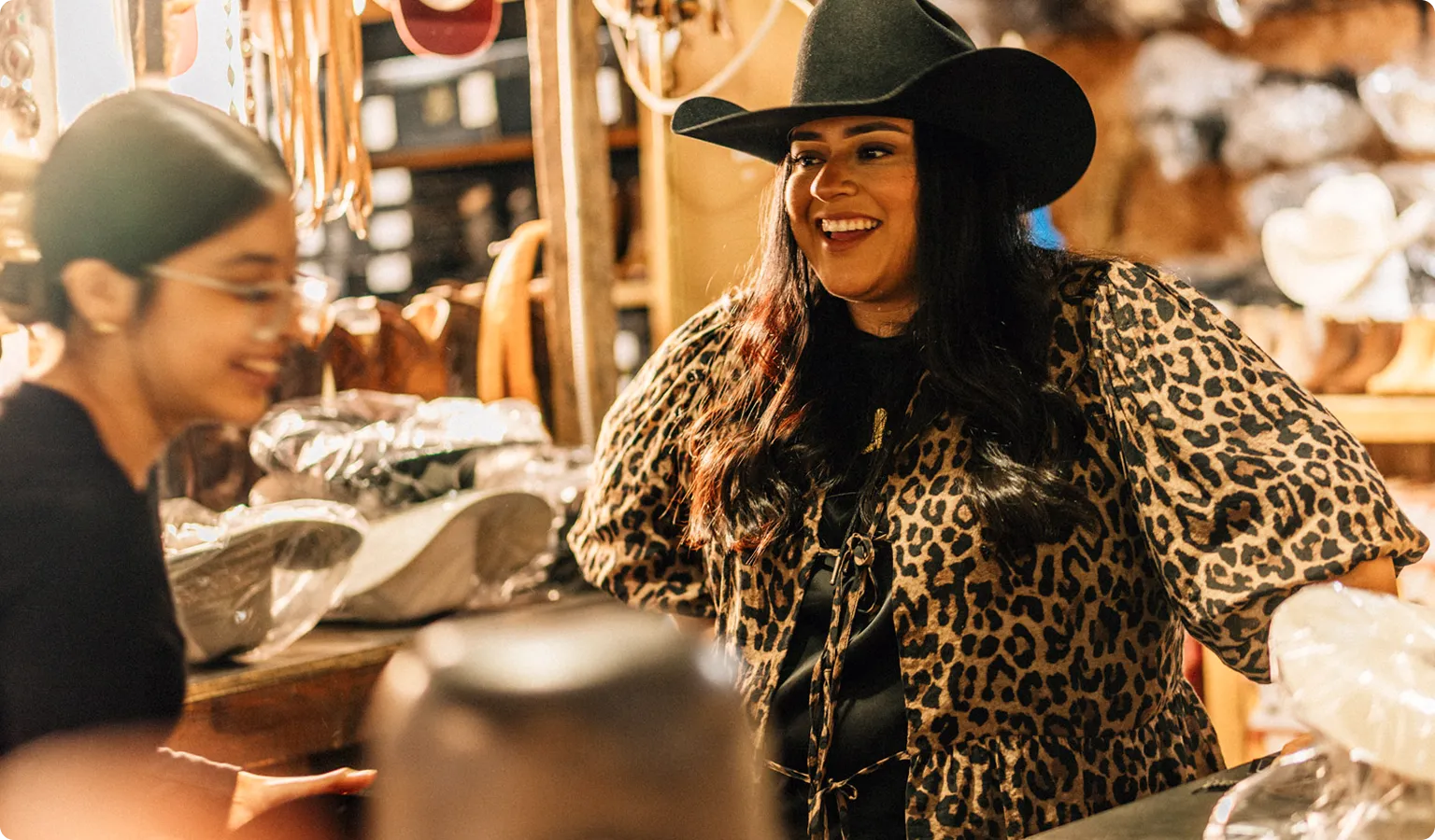 Woman wearing a black cowboy hat and leopard print blouse smiling and talking with another woman in a rustic shop.