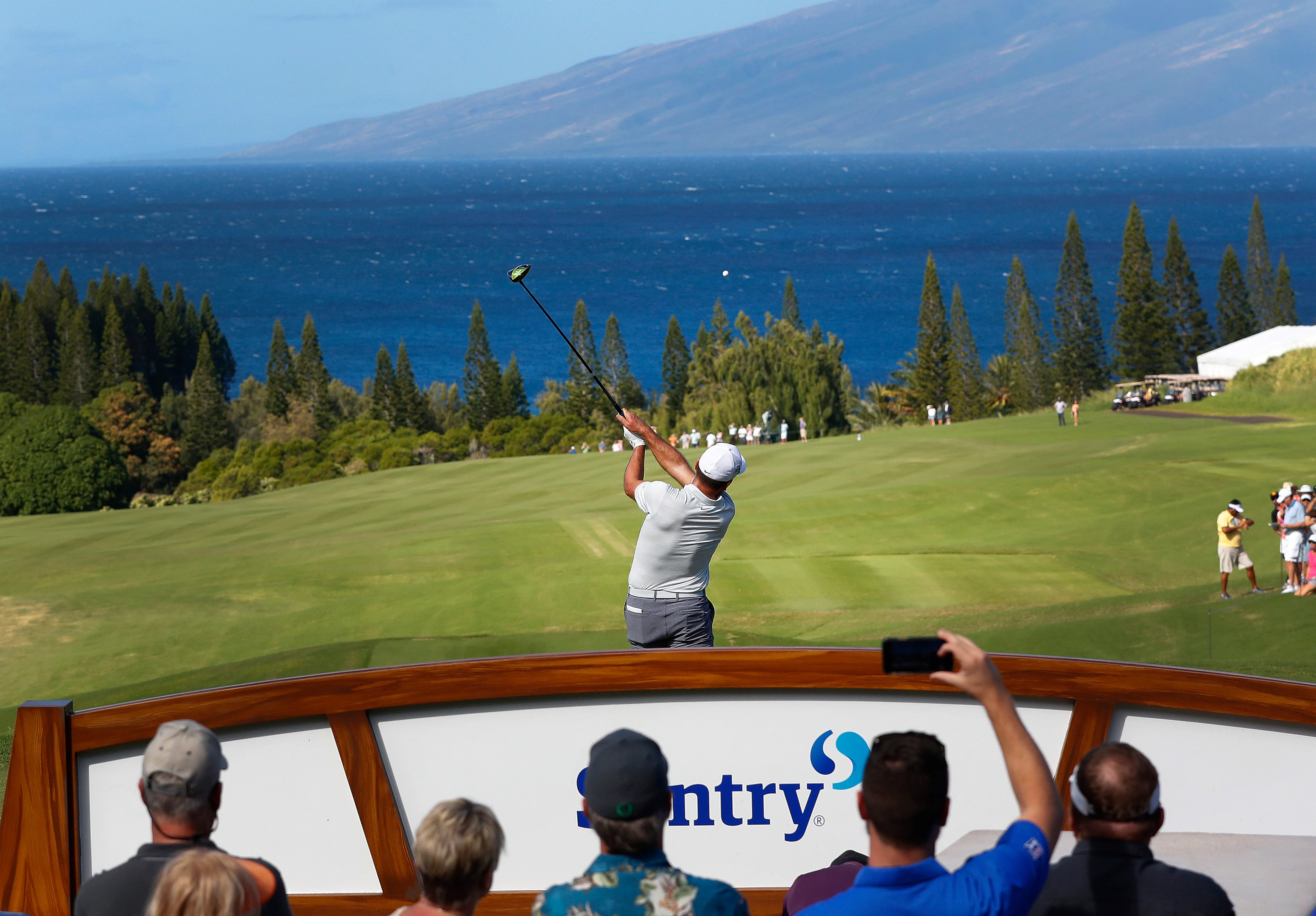Golfer in mid-swing on a lush green course with spectators watching and ocean and mountains in the background.