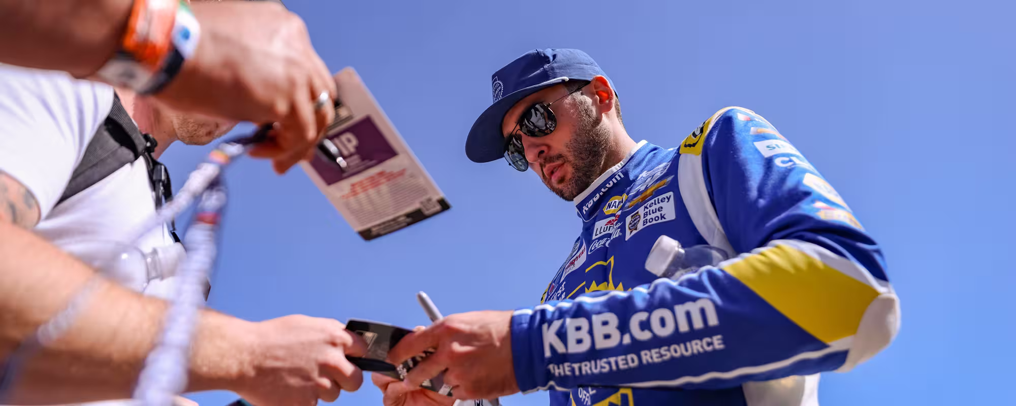 Race car driver Chase Elliott in blue KBB.com racing suit signing autographs for fans.