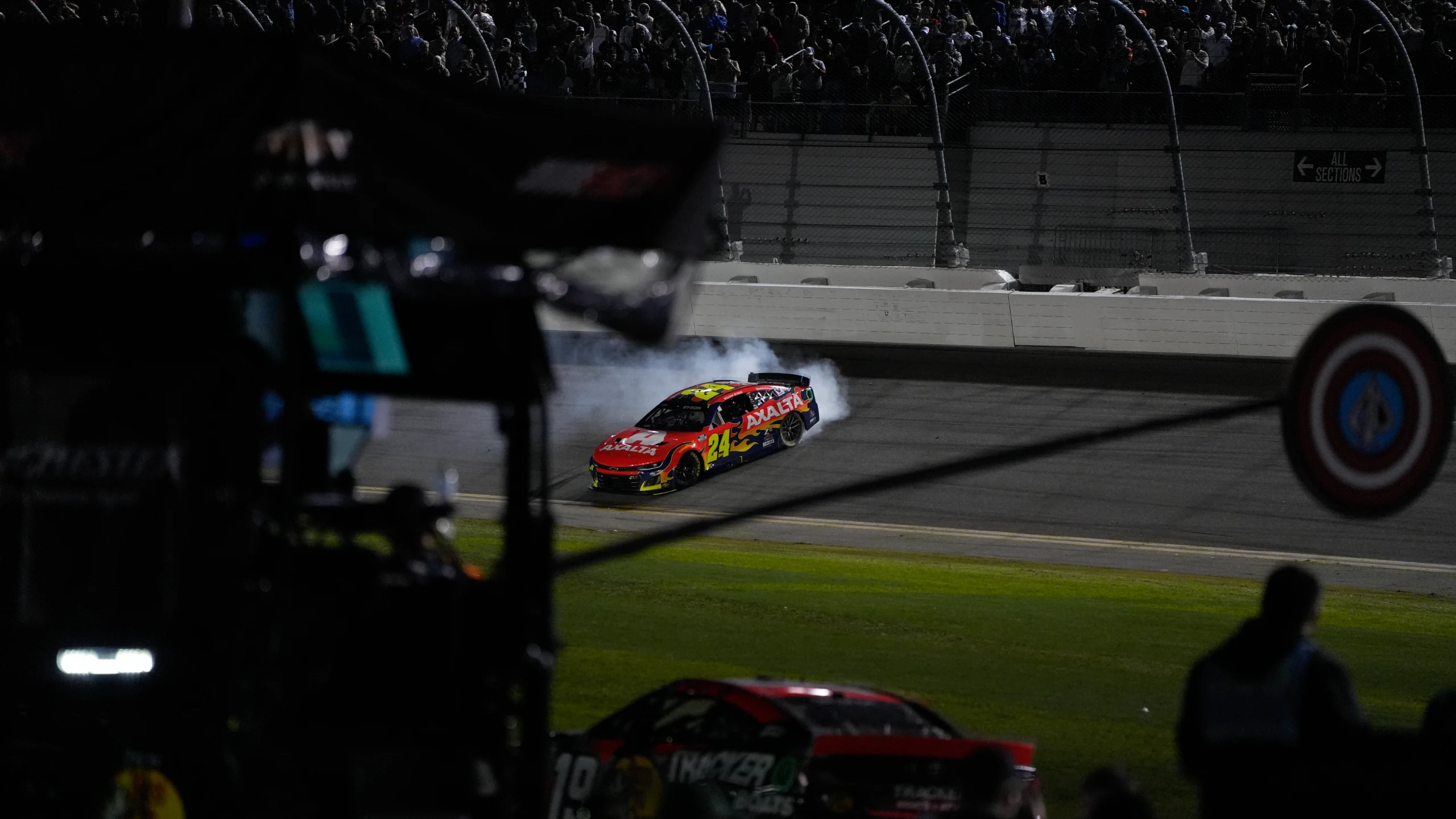 Race car number 24 with Axalta branding skidding and producing smoke on a race track at night with spectators in the stands.