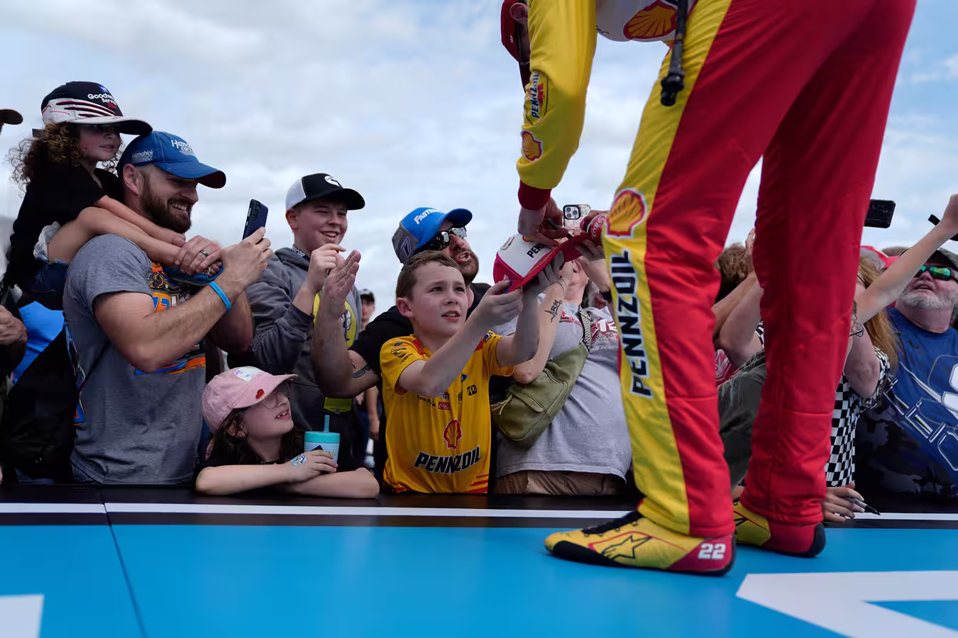 Crowd of fans, including children, excitedly reaching out to a race car driver in red and yellow Pennzoil racing gear to get an autograph and take photos.