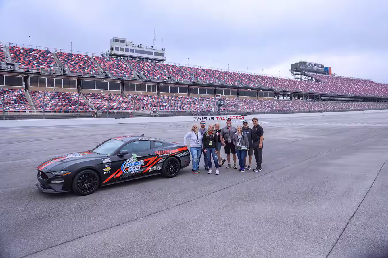 Group of seven people standing beside a black and orange GEICO 500 race car on the Talladega Superspeedway race track with empty grandstands in the background.