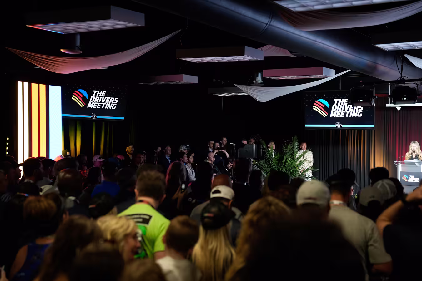 Crowd of people attending The Drivers Meeting event with speaker at podium and event logo on screens.