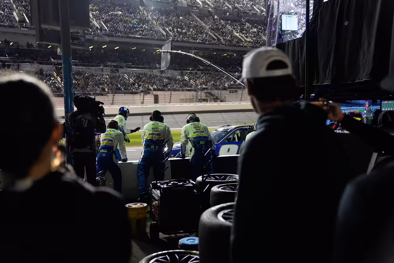 Pit crew members in yellow and blue uniforms standing by a race car at night during a NASCAR event with a large crowd in the stands.