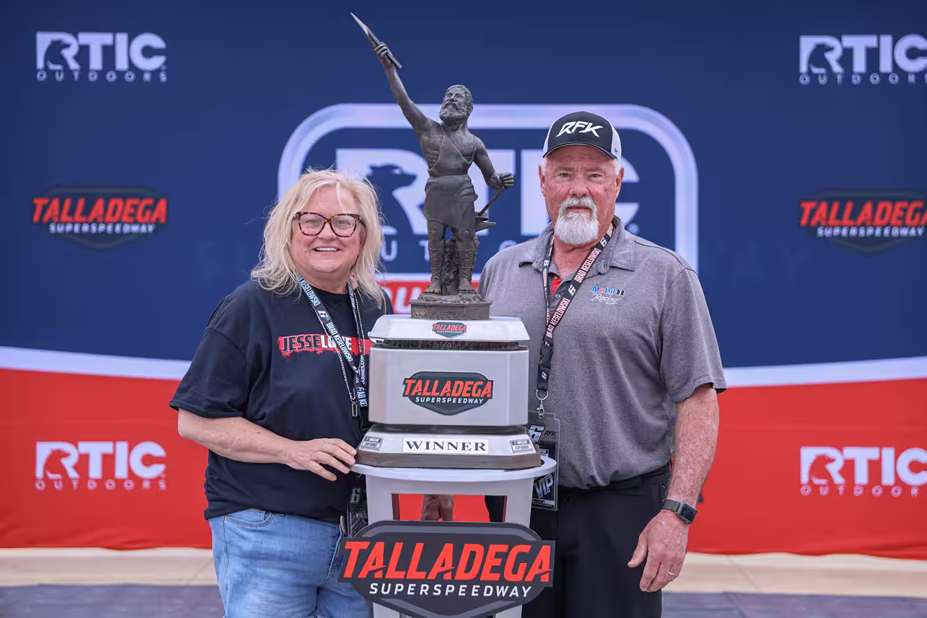 Two people standing beside the Talladega Superspeedway winner trophy with a statue holding a spear.