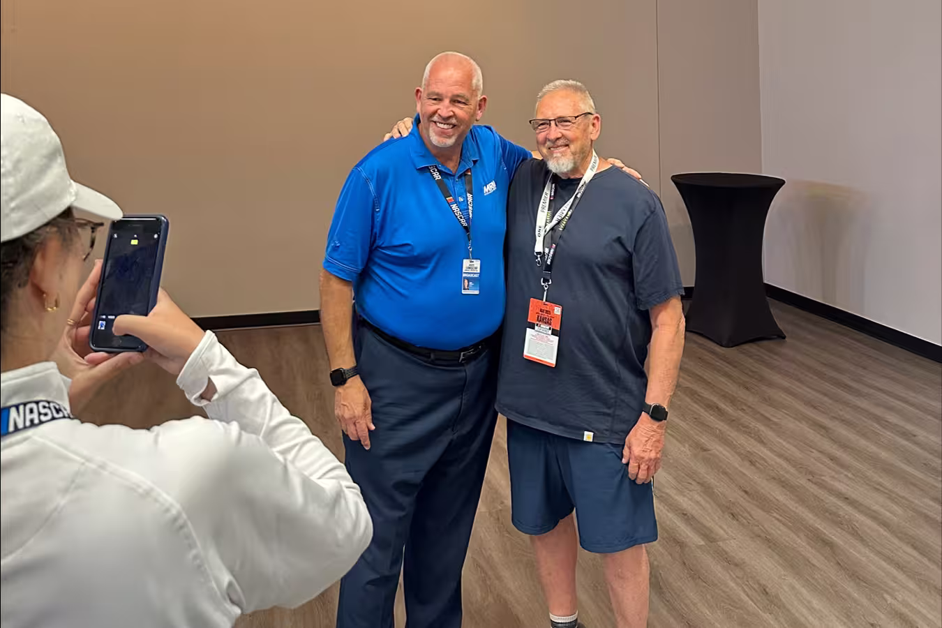 Two smiling older men posing for a photo indoors, one wearing a blue polo shirt and the other in a dark t-shirt and shorts, while a woman in a white cap takes their picture with a smartphone.