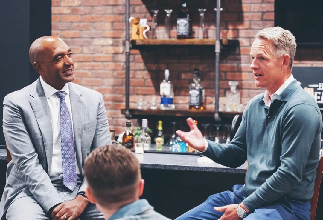 Three men engaged in a conversation in a room with brick walls and shelves holding bottles and glassware.
