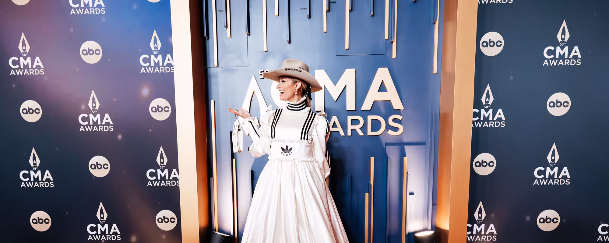 Woman in a white dress with black stripes and a beige cowboy hat posed on the red carpet at the CMA Awards.