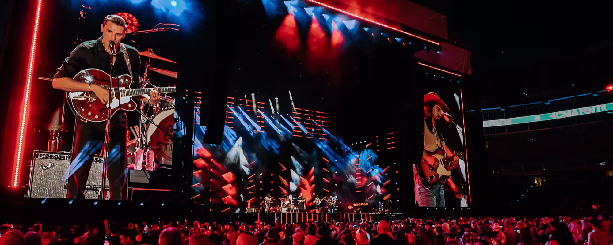 Crowd watching a live band performing on a brightly lit stage with large screens displaying musicians playing guitars.
