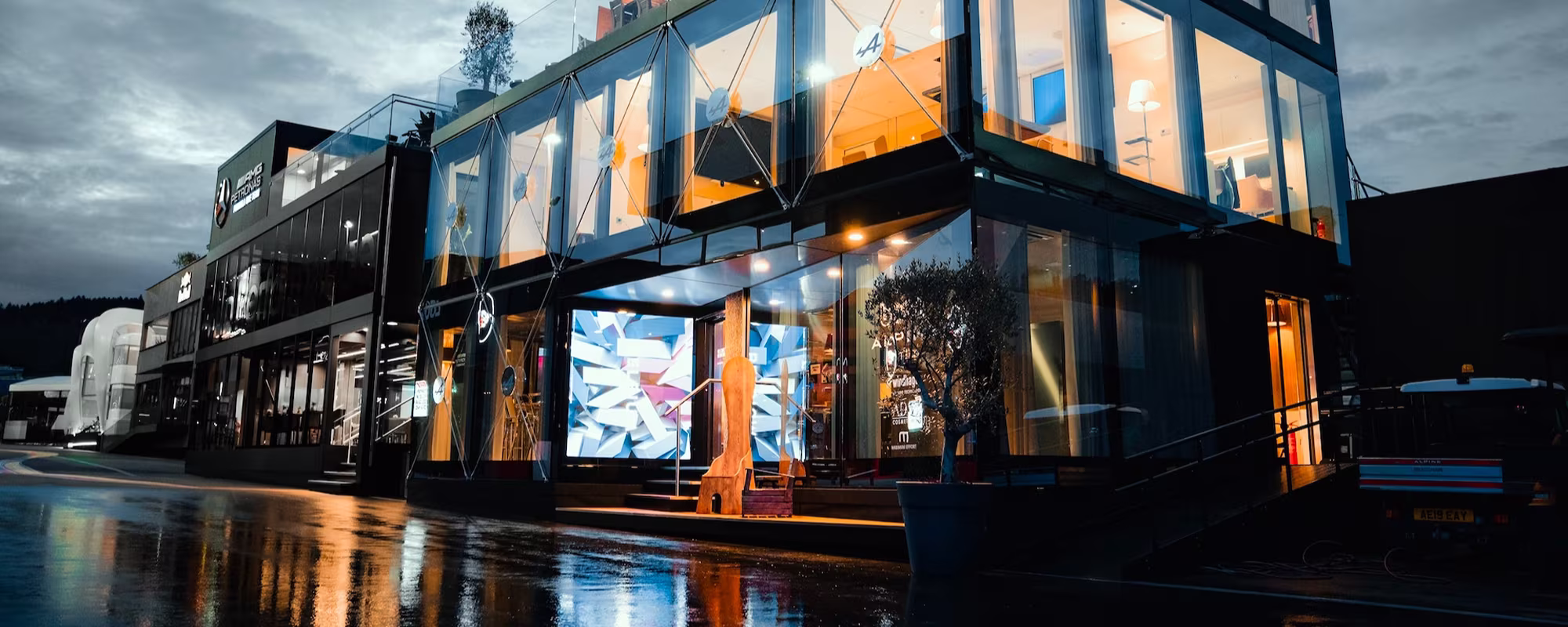 Modern two-story glass building at dusk with warm interior lights reflecting on wet pavement.