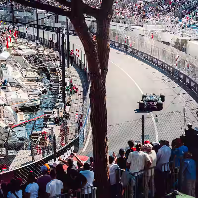 Formula 1 race car driving on a Monaco street circuit with yachts and spectators lining the track.