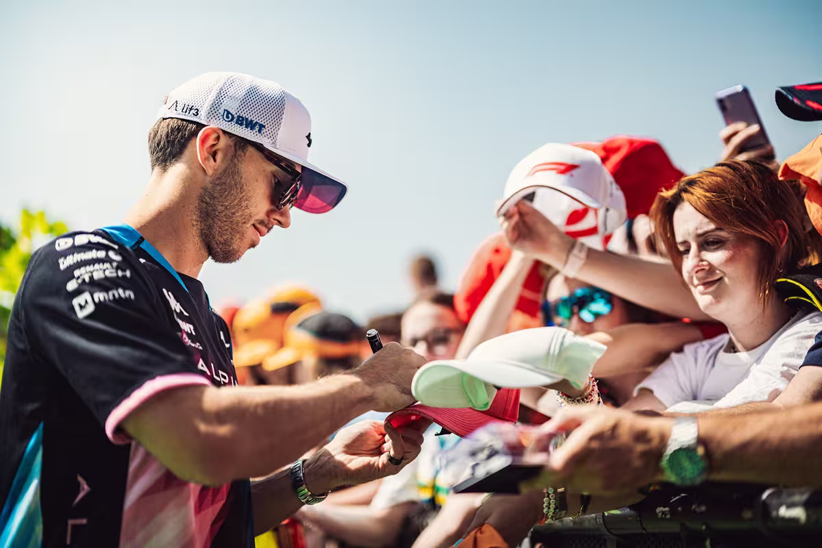 Male race car driver signing a red cap for fans behind a barrier on a sunny day.