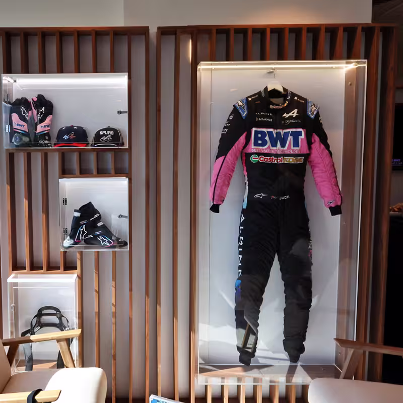 Display of a black and pink racing suit with BWT and Castrol logos, gloves, hats, and racing shoes in a wood-paneled room.