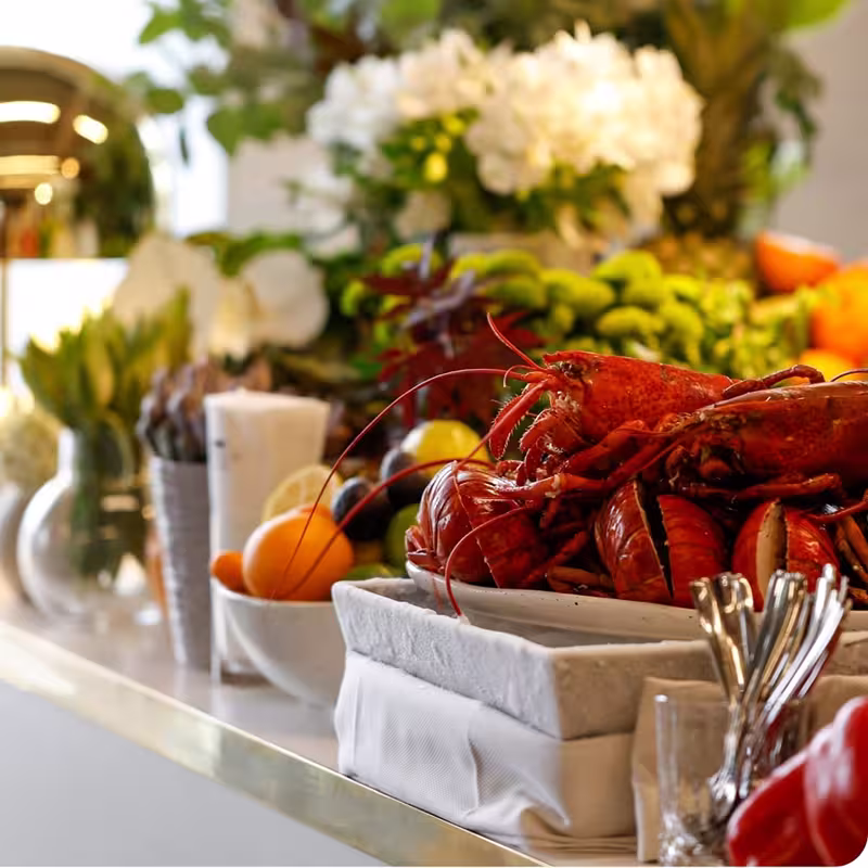 Plate of cooked lobsters on a white table with bowls of fruit and out-of-focus flowers in the background.