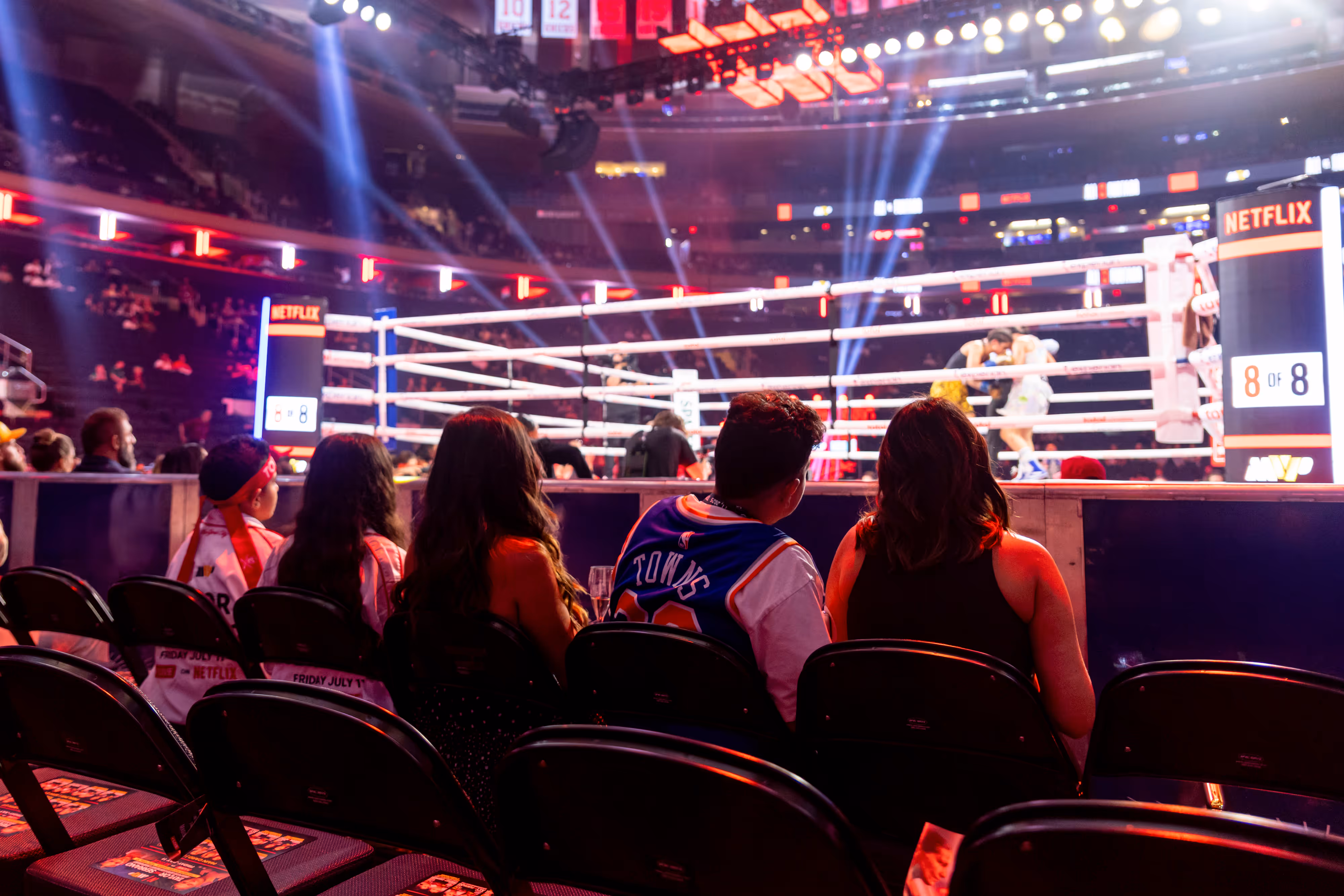 Four spectators sitting front row watching a boxing match in a brightly lit arena with ringside seats and Netflix branding.