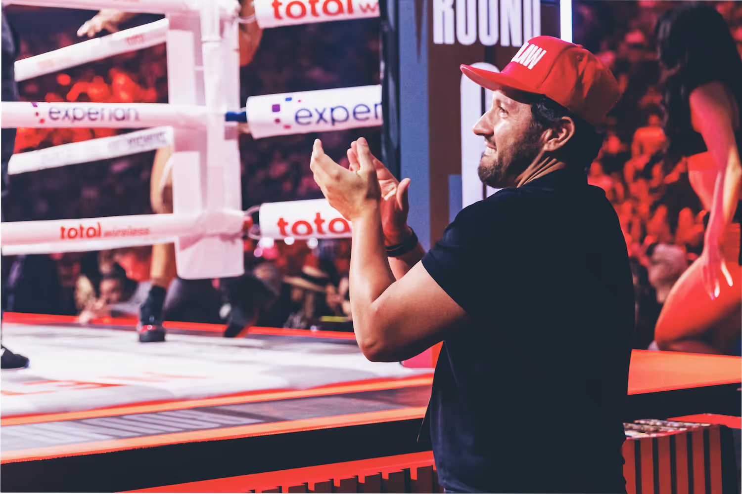 Man in a red cap clapping near a boxing ring during a match.