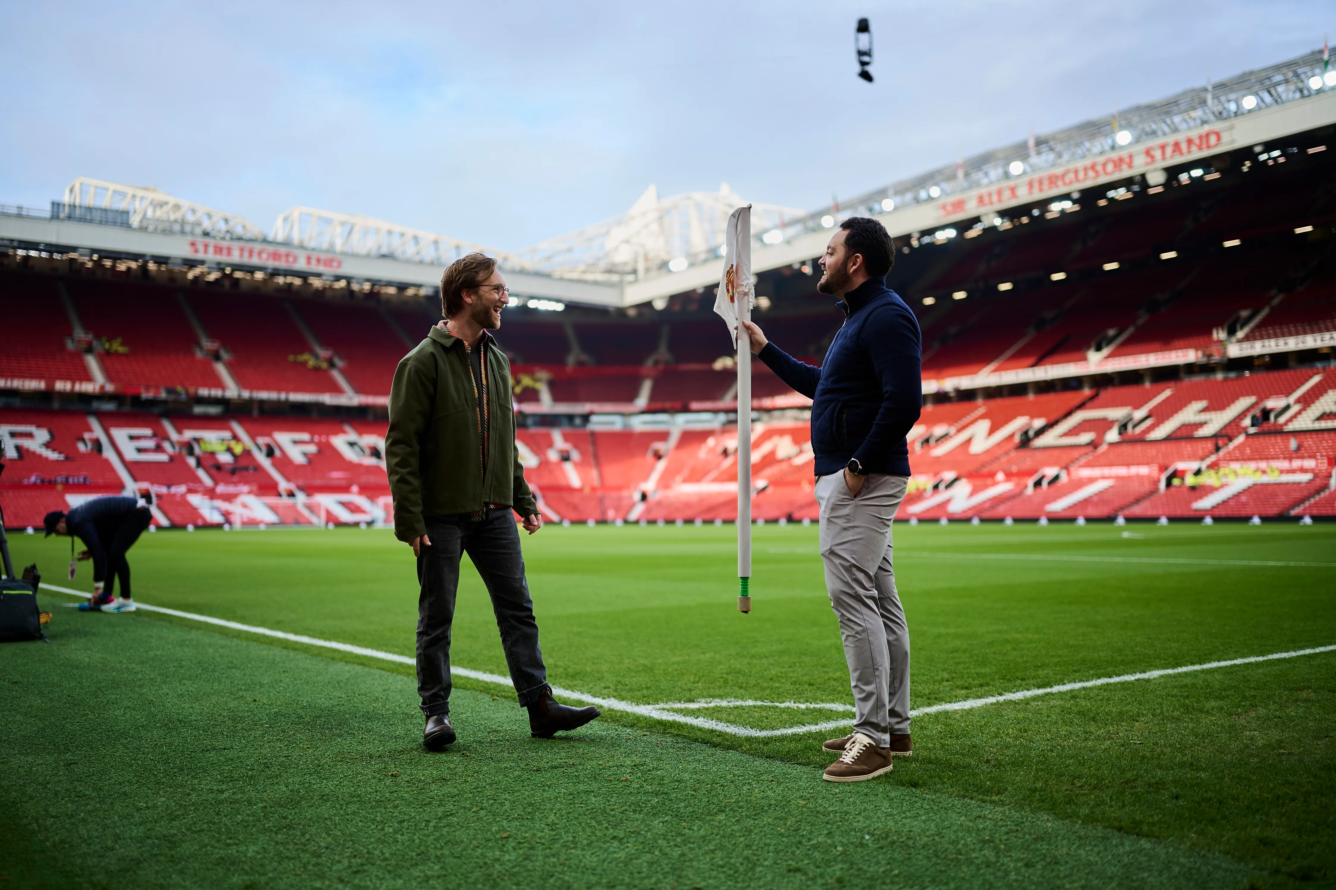 Two men standing and talking near the corner flag on a football pitch inside an empty stadium.