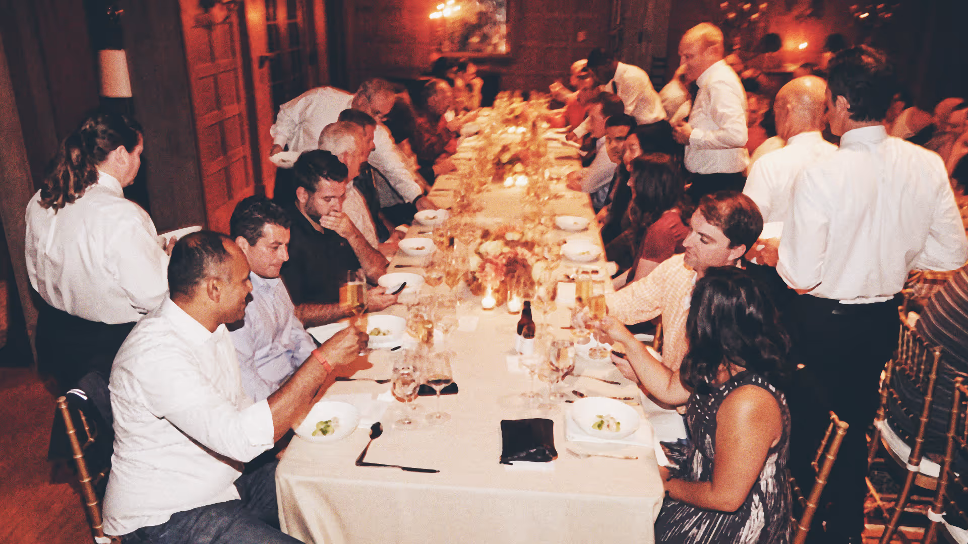 People seated at a long, candlelit dining table in a warmly lit room, some raising glasses in a toast while servers attend to them.