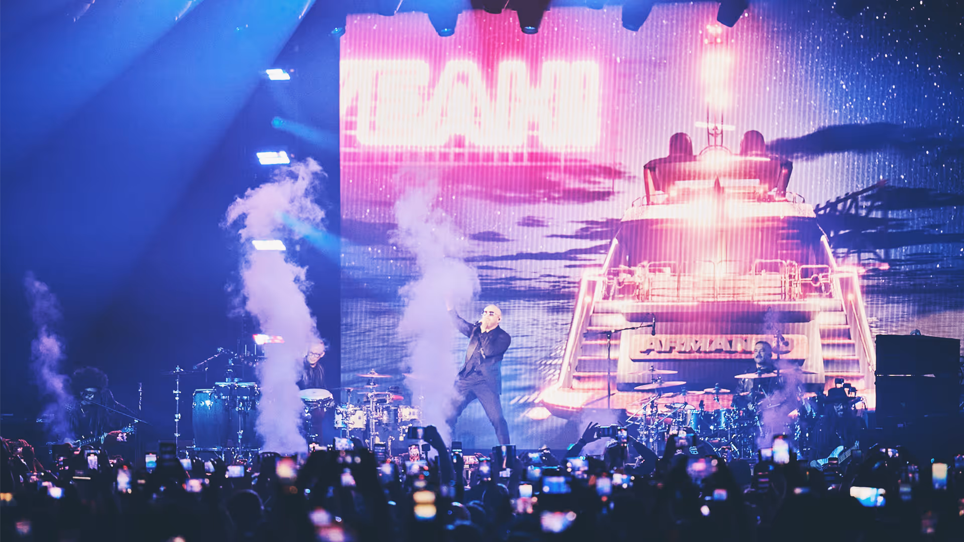 Live concert with a singer performing on stage surrounded by musicians and smoke effects, with an audience holding up phones capturing the event.