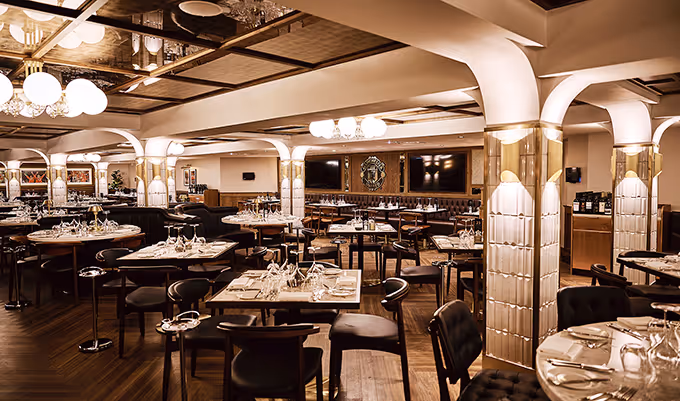 Elegant restaurant interior with dark wood floors, round and square tables set with wine glasses and cutlery, and decorative columns with gold accents.