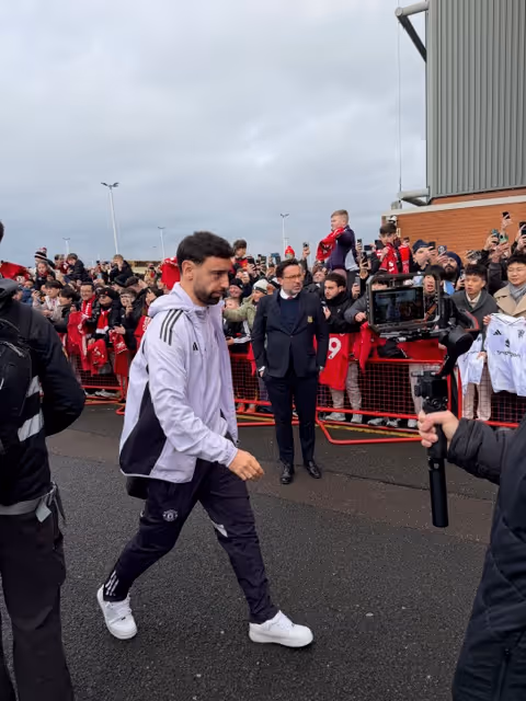 Soccer player in white jacket and black pants walking past excited fans behind a barrier at an outdoor event.