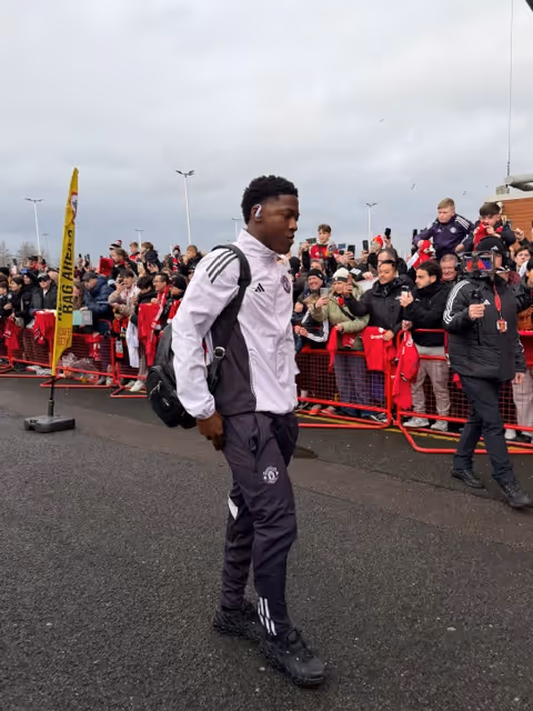 Soccer player in Manchester United tracksuit walking past fans taking photos behind a red barricade.