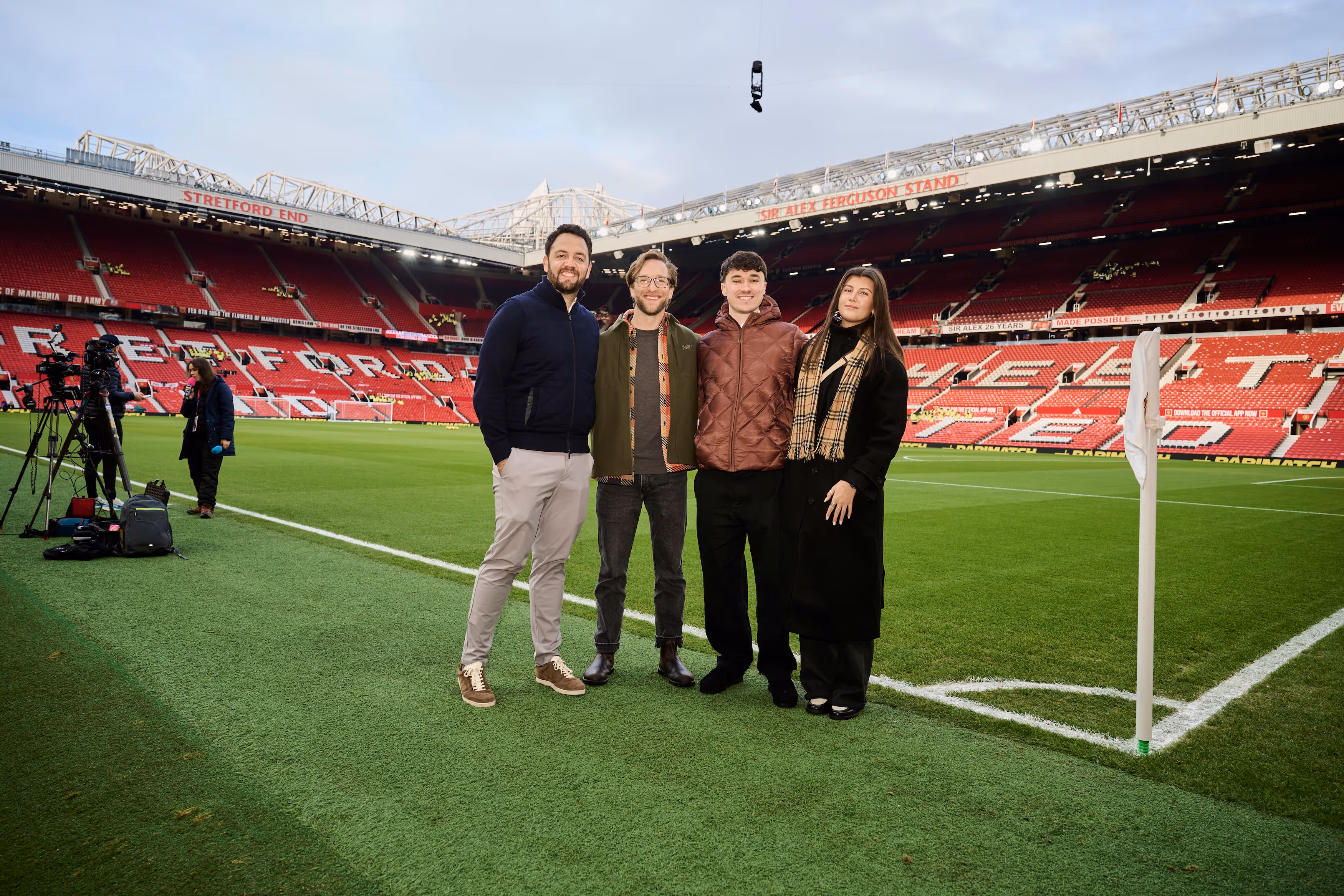 Four people standing side by side on the grass near a corner flag inside a large football stadium with empty red seats and 'STRETFORD END' and 'SIR ALEX FERGUSON STAND' signs visible.