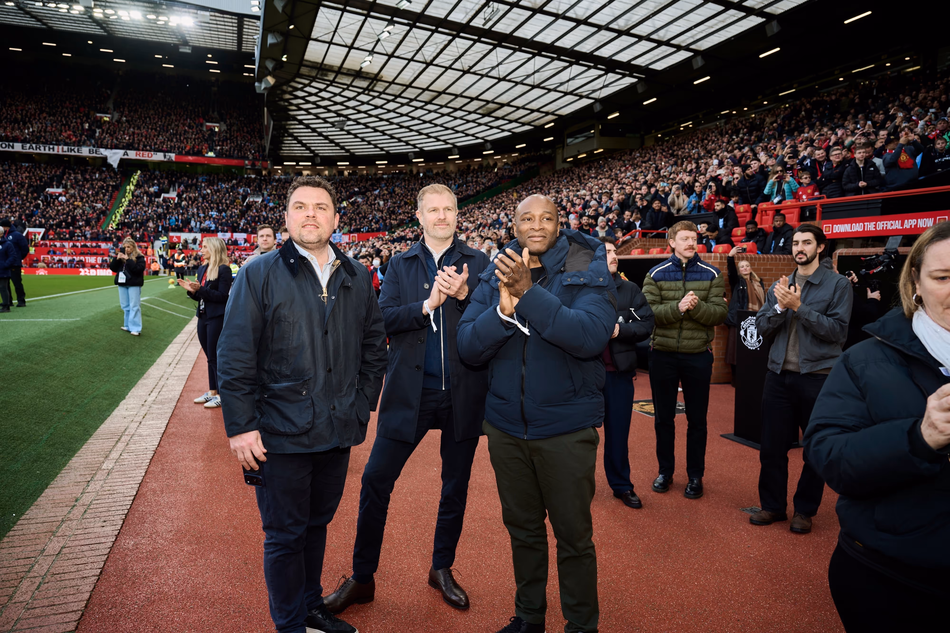 Group of men standing and applauding on a stadium track with a large crowd in the stands behind them.