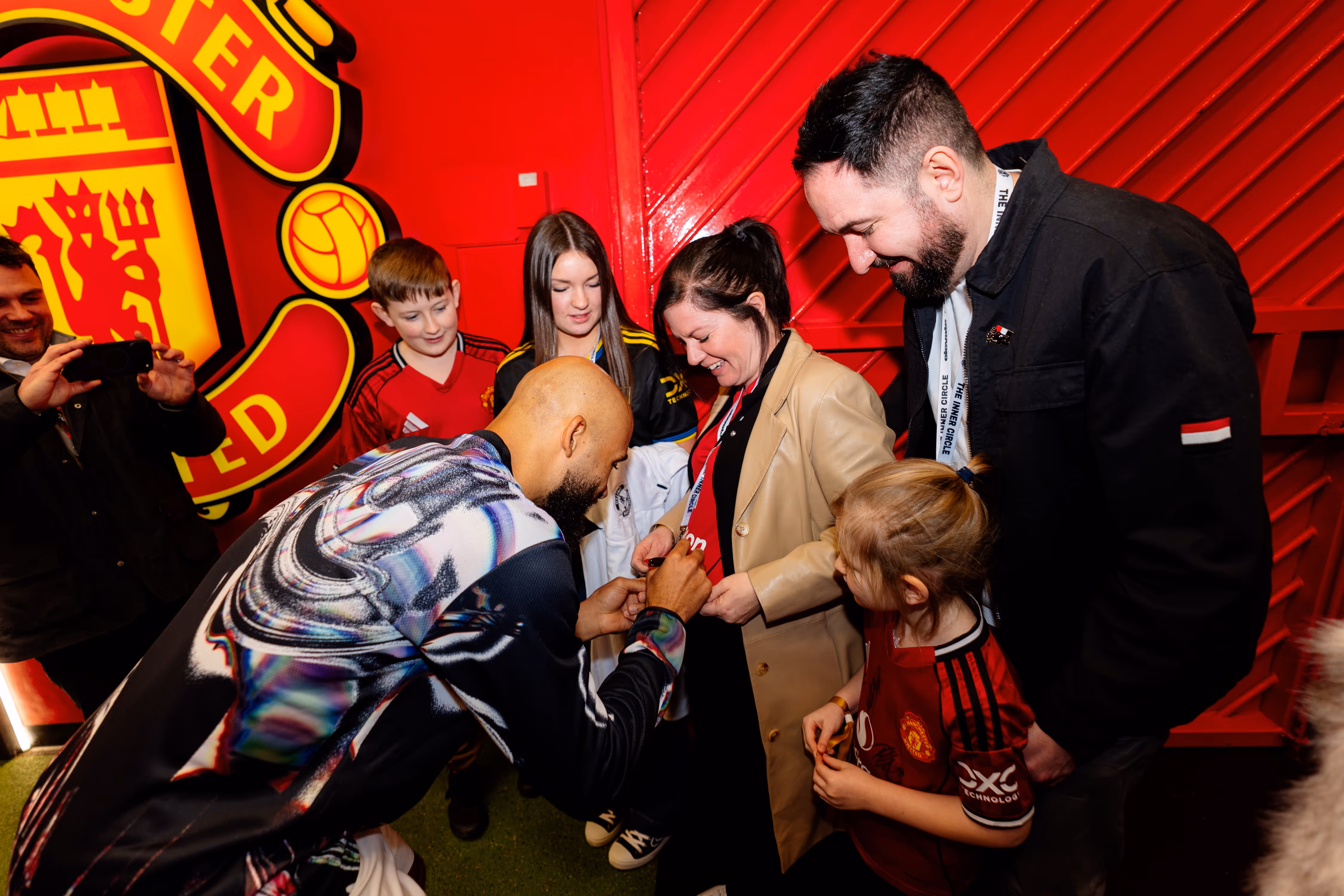 Football player signing a fan's shirt surrounded by smiling children and adults in Manchester United apparel under a large Manchester United logo.
