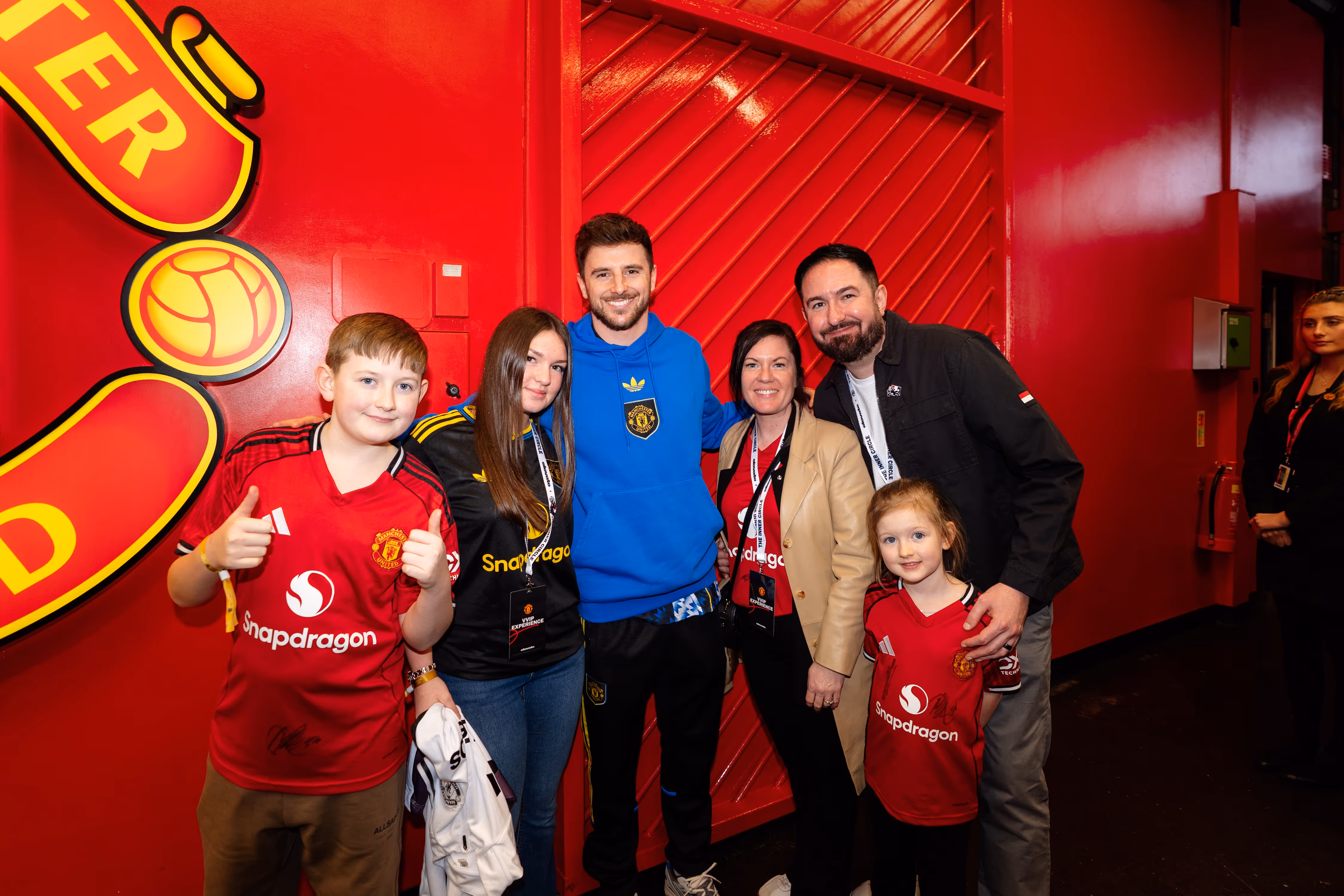 Group of five smiling people including children wearing Manchester United jerseys posing in front of a large red wall with part of the Manchester United logo visible.