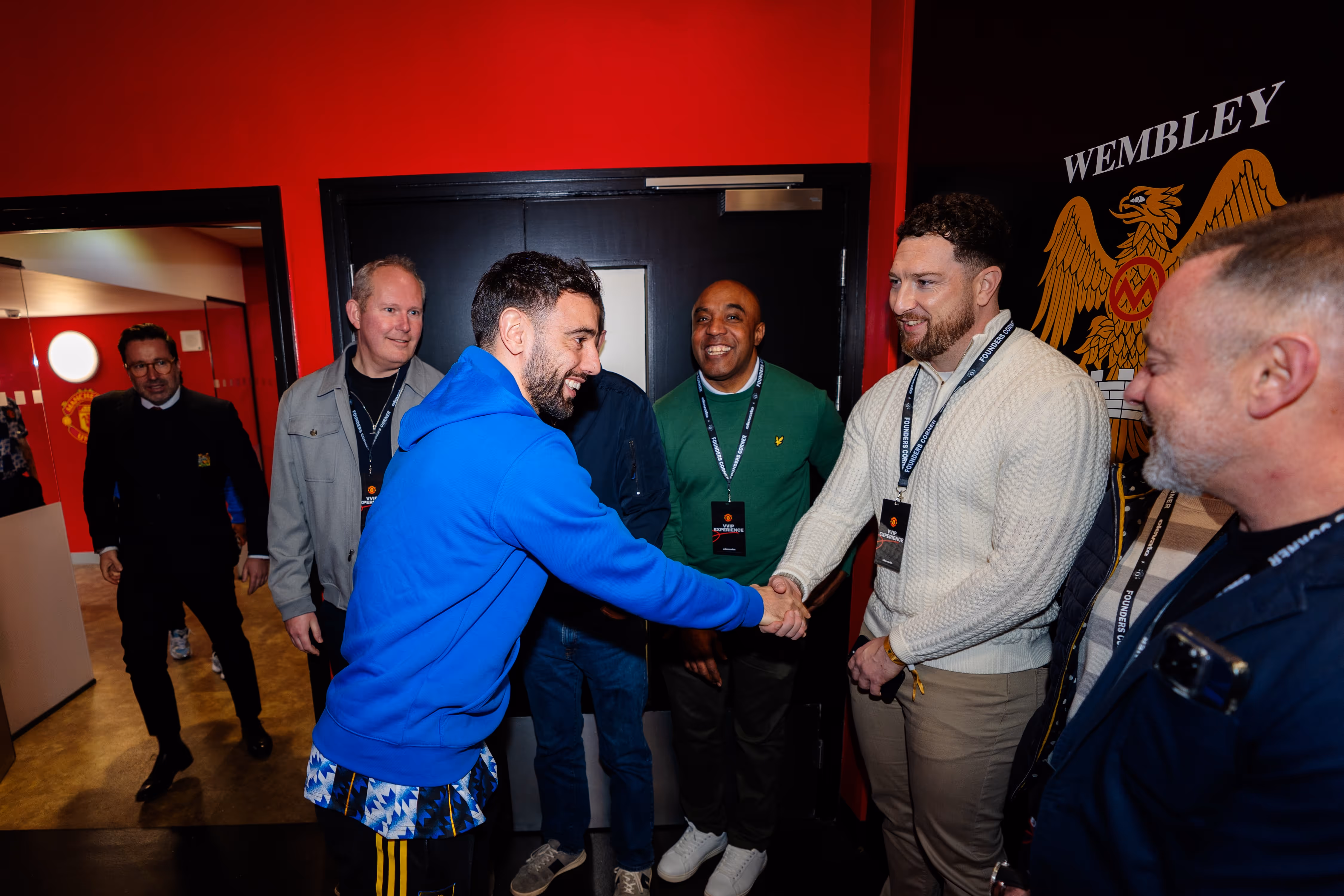 A man in a blue hoodie shaking hands with another man wearing a white sweater, surrounded by smiling people inside a venue with red and black walls and a Wembley emblem.
