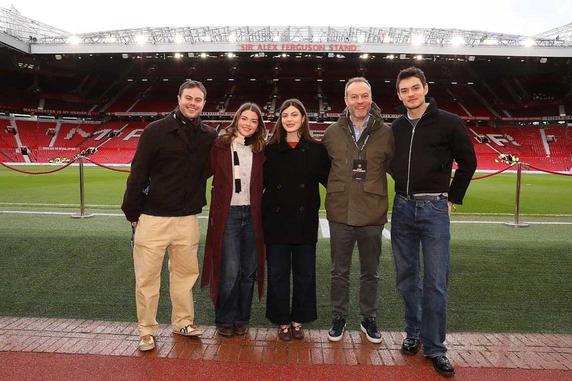 Five people standing together on the edge of a football pitch inside Old Trafford stadium, with red seats and the Sir Alex Ferguson Stand visible in the background.
