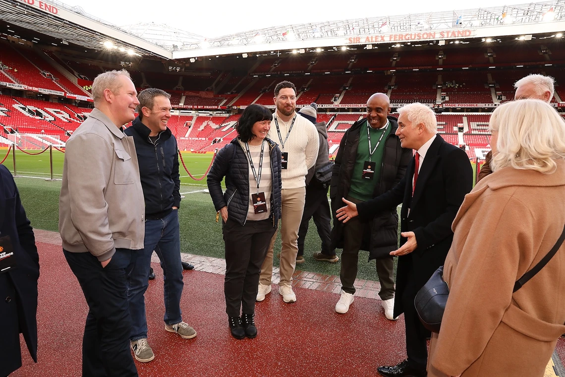 A group of people smiling and talking on the edge of a football pitch inside a stadium with red seats and the Sir Alex Ferguson Stand in the background.