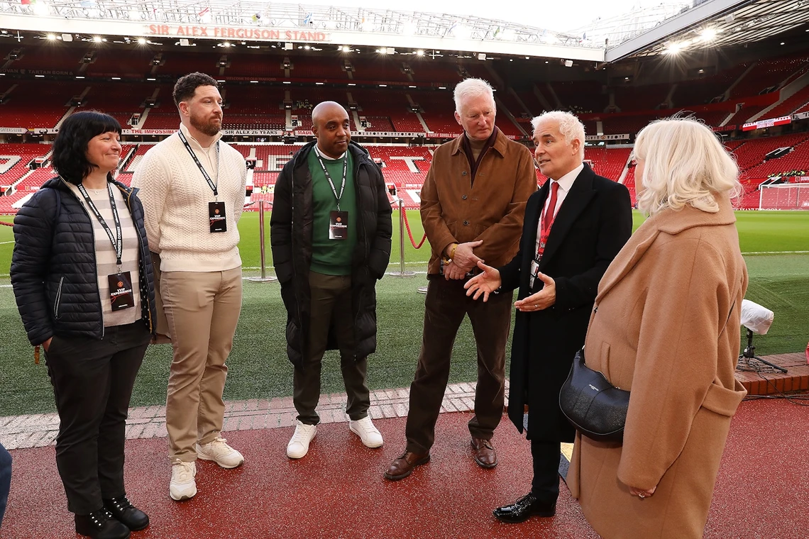 Six people standing on the edge of a football pitch inside a stadium, having a conversation.