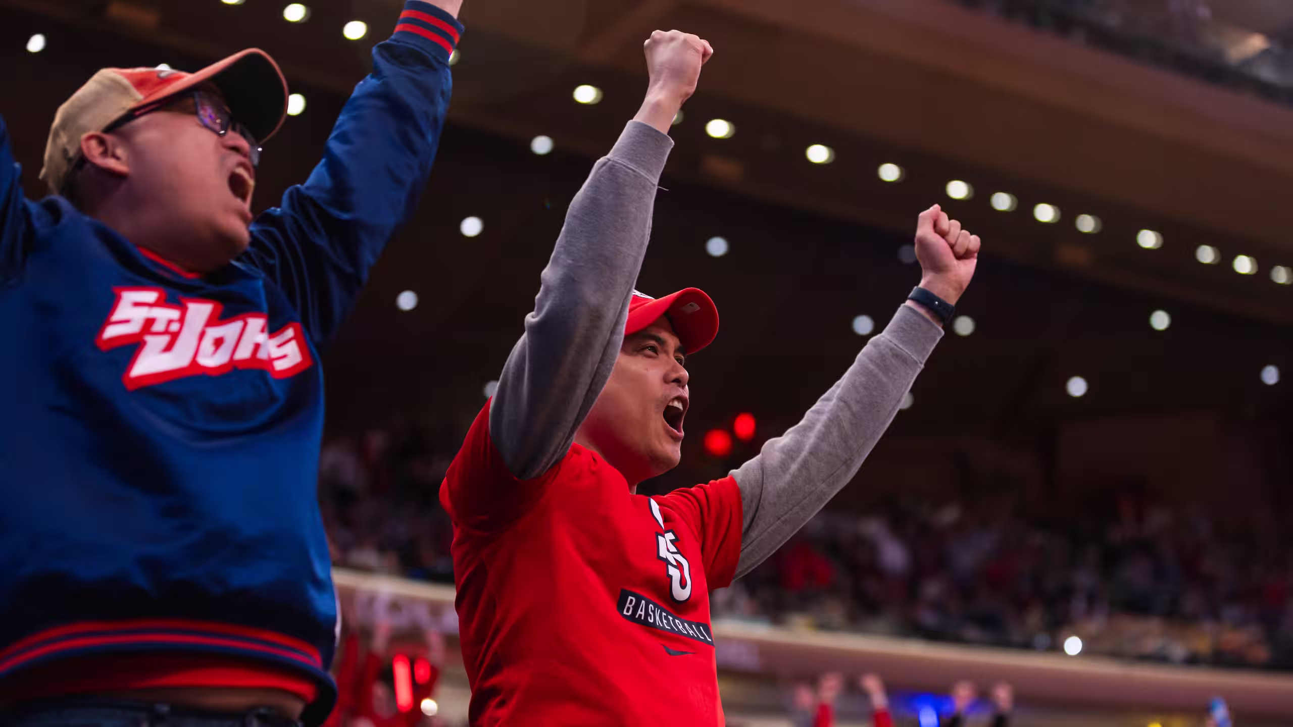 Two enthusiastic fans cheering with raised fists at a basketball game in an indoor stadium.