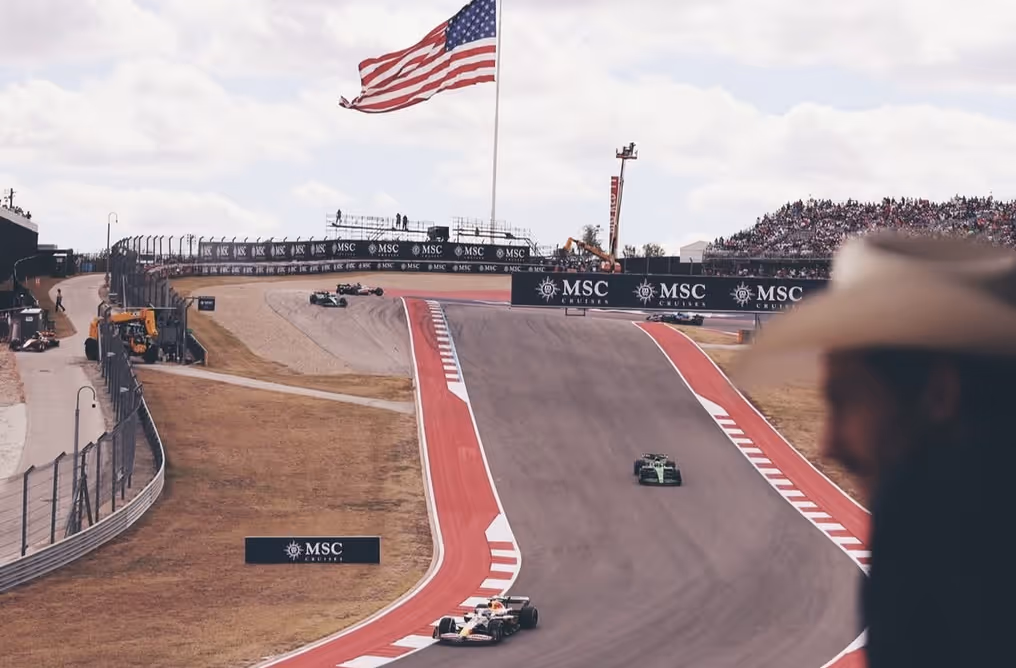 Formula 1 cars racing on a track with red and white curbs, a large American flag flying, and spectators in grandstands.