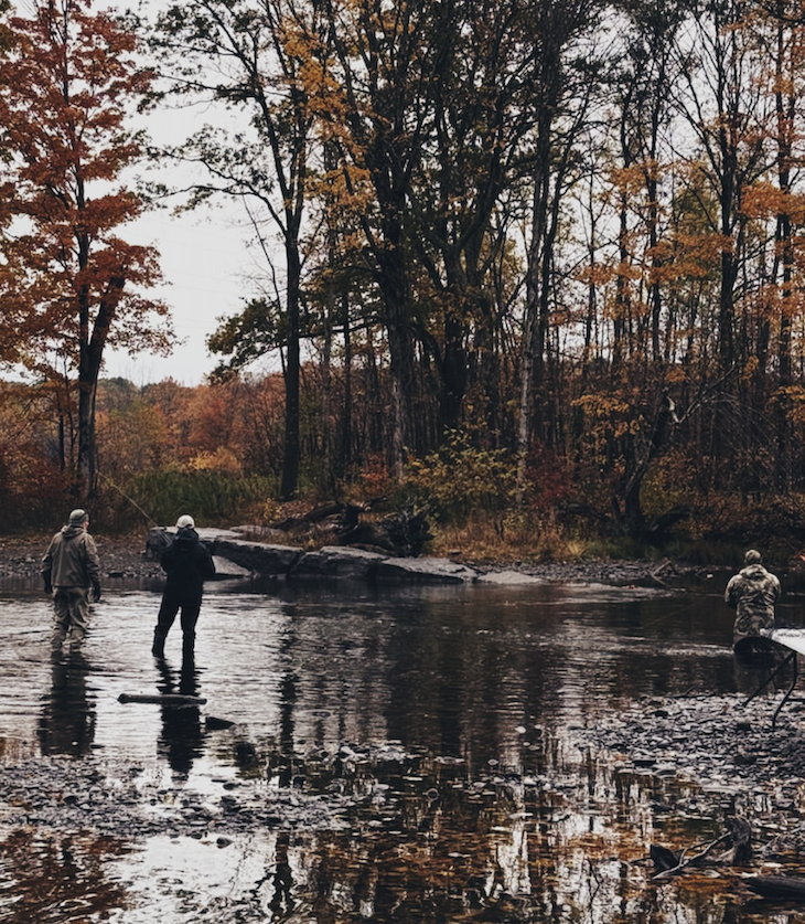 Three people fishing in a shallow river surrounded by trees with autumn foliage.