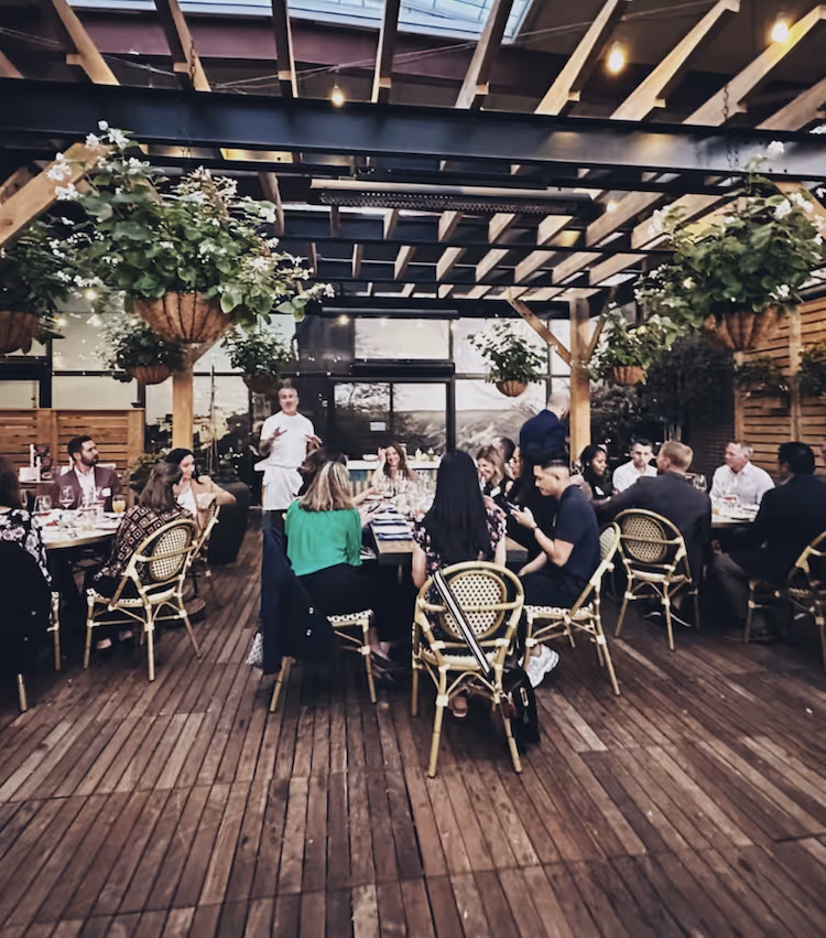 Group of people dining at a long table under a wooden pergola with hanging plants in an outdoor restaurant.