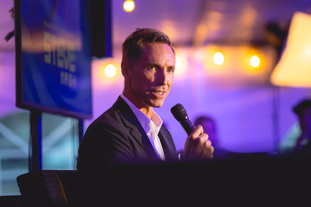 Man in a dark suit speaking into a microphone at an event with purple lighting and blurred background lights.