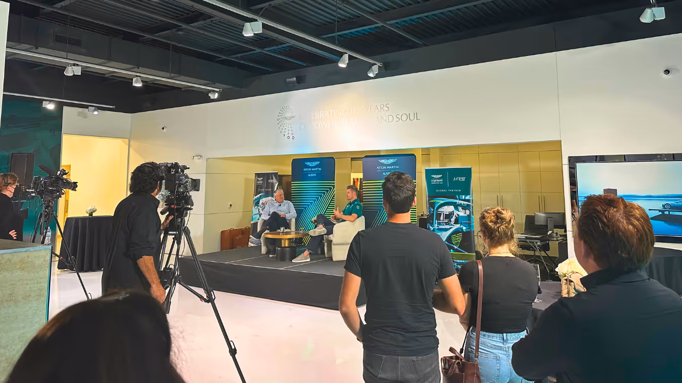 Audience watching a recorded interview or talk show on a stage with two men seated, cameras filming the event in a modern indoor setting.
