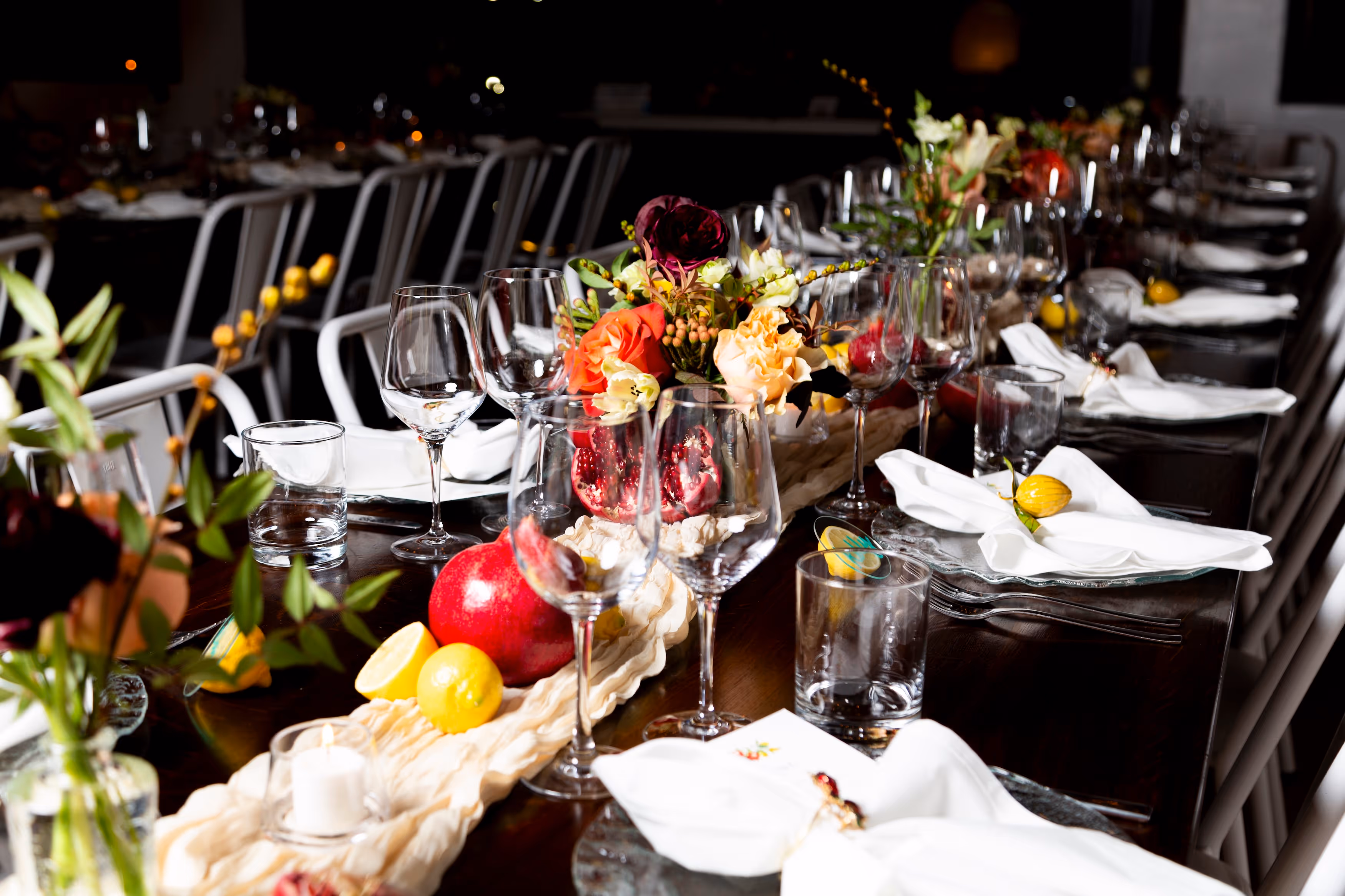Long dining table with white chairs, decorated with colorful flowers, pomegranates, lemons, wine glasses, and white napkins.